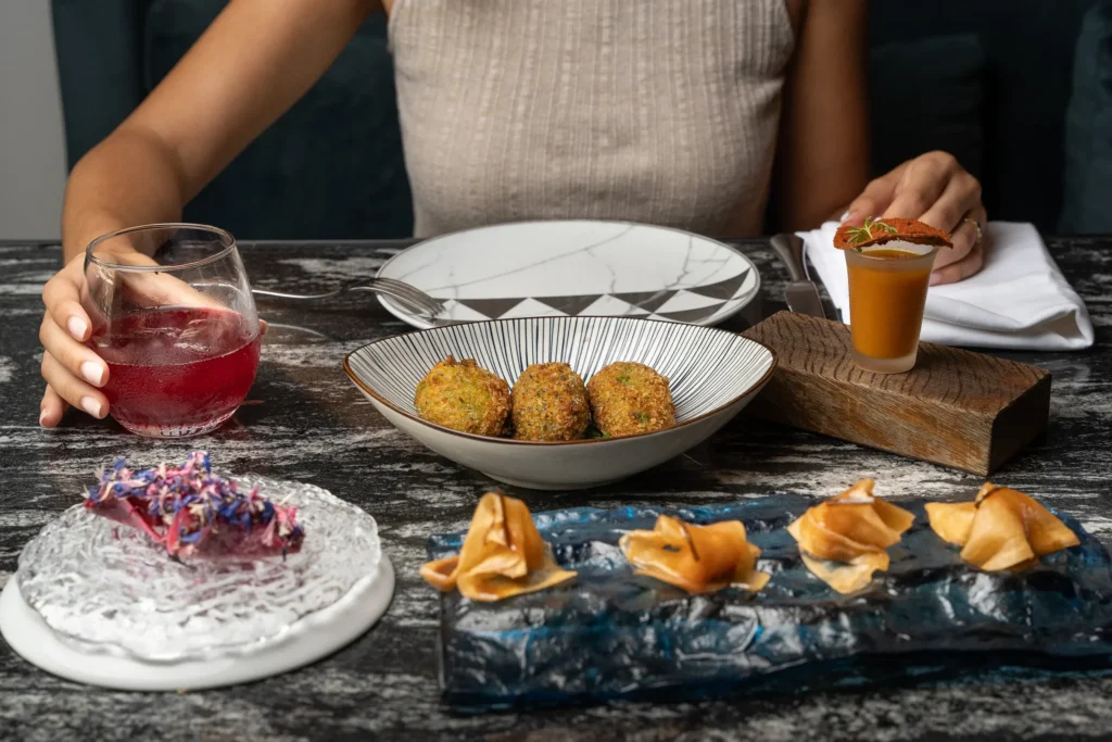 A person sits at a table with various plated appetizers, a red beverage in a glass, and a napkin with utensils.